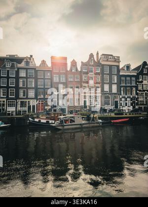 Vue sur le canal reflétant les façades vibrantes des bâtiments historiques sous un ciel nuageux, ponctué par le soleil, Amsterdam-centrum, Noord-Holl Banque D'Images