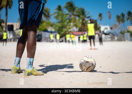 PAJE, ZANZIBAR - 18 OCTOBRE 2025 : gros plan des jambes d'un joueur de football et d'un ballon sur une plage de sable fin lors d'une séance d'entraînement avec des palmiers dans le b Banque D'Images