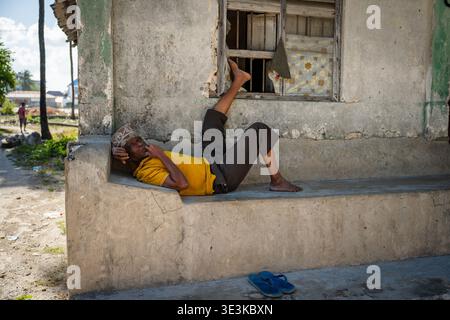 PAJE, ZANZIBAR - 18 OCTOBRE 2025 : un homme du coin se détendant sur un banc de pierre à l'extérieur d'un bâtiment traditionnel à Zanzibar, reflétant le calme et la lenteur Banque D'Images