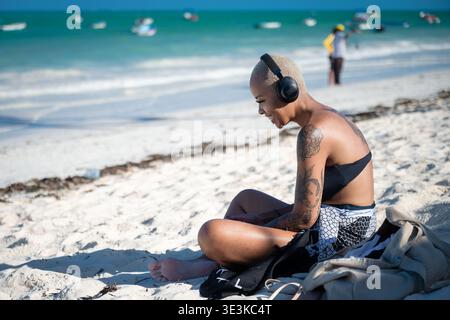 PAJE, ZANZIBAR - 18 OCTOBRE 2025 : une jeune femme tatouée assise sur une plage de sable blanc, portant des écouteurs et profitant de la musique sur fond Banque D'Images