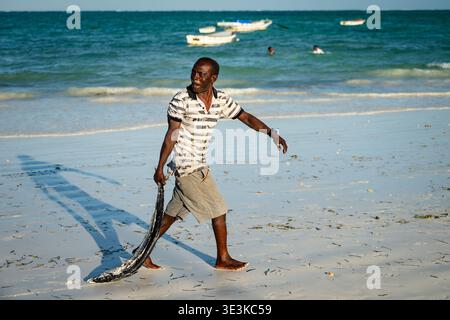 PAJE, ZANZIBAR - 18 OCTOBRE 2025 : un pêcheur local marchant le long de la plage tropicale de Zanzibar, transportant un gros poisson pêché dans l'eau turquoise Banque D'Images