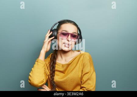 Une femme se tient contre un mur solide tout en portant des écouteurs et des lunettes de soleil. Elle aime la musique et montre une expression détendue. Ses cheveux sont tressés et Banque D'Images