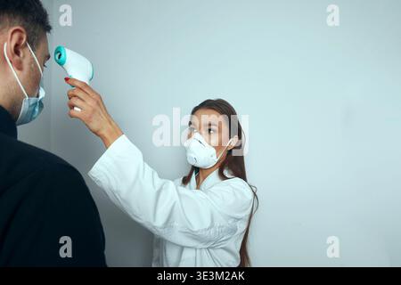 Un agent de santé vérifie la température d'un patient avec un thermomètre numérique. Les deux portent un masque facial. La scène se déroule dans un établissement médical Banque D'Images