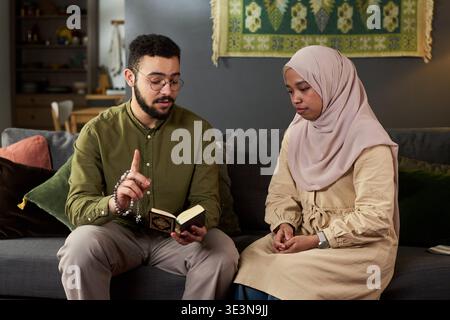 Jeune homme musulman adulte expliquant un texte religieux à une jeune femme adulte portant le hijab alors qu'elle était assise sur un canapé, les deux se concentrant sur le livre, l'homme faisant des gestes avec la main, la femme écoutant attentivement Banque D'Images