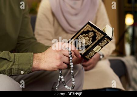 Jeune homme musulman tenant le Coran et des perles de prière assis à côté d'une jeune femme musulmane adulte portant le hijab, tous deux participant à la pratique religieuse dans un cadre intérieur Banque D'Images