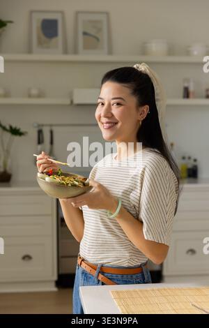 Femme asiatique portant un tee-shirt rayé et un Jean souriant tenant un bol en céramique à l'aide de baguettes dans la cuisine Banque D'Images