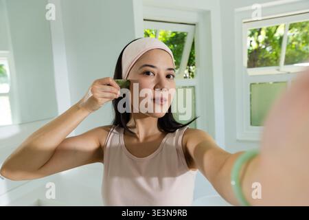 Femme afro-américaine en haut rose pâle, bandeau appliquant le jade gua Sha, tenant le téléphone dans la salle de bain Banque D'Images