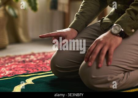 Jeune homme musulman agenouillé sur un tapis de prière levant le doigt pendant le rituel de prière islamique, les mains reposant sur les cuisses, portant une montre-bracelet, pratiquant la pratique religieuse à l'intérieur Banque D'Images