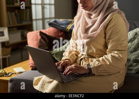 Femme caucasienne musulmane d'âge moyen assise sur un canapé à l'aide d'un ordinateur portable, portant un foulard, se concentrant sur la frappe, le travail ou l'étude dans un environnement domestique, les mains sur le clavier Banque D'Images