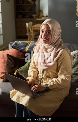 Portrait de jeune femme musulmane adulte souriant tout en étant assise sur un canapé à l'aide d'un ordinateur portable, les mains sur le clavier, portant le hijab, regardant dans la caméra dans le cadre du salon à la maison Banque D'Images
