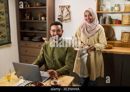 Jeune homme adulte moyen-oriental assis à table à l'aide d'un ordinateur portable et souriant tandis que jeune femme adulte moyen-orientale debout à côté de lui, tenant une serviette de vaisselle et souriant dans le cadre de la cuisine Banque D'Images
