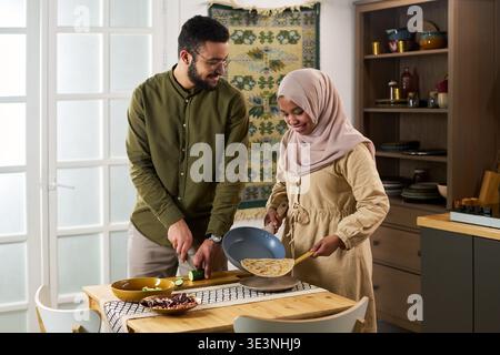 Jeune homme musulman adulte et jeune femme musulmane adulte préparant la nourriture ensemble dans la cuisine, l'homme tranchant les légumes tandis que la femme tenant la poêle à frire avec du pain plat Banque D'Images