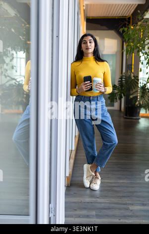 Femme indienne appuyée contre la cloison en verre tenant la tasse à café smartphone dans le couloir du bureau Banque D'Images