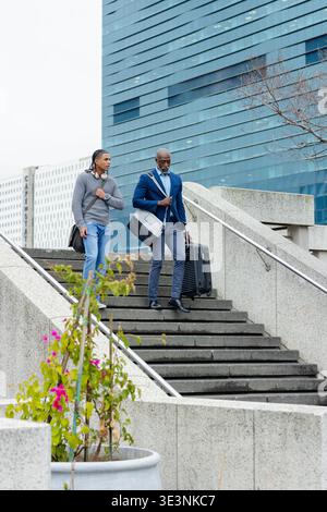 Divers amis masculins marchant dans les escaliers de la plaza en béton transportant valise roulante et sac de sport Banque D'Images
