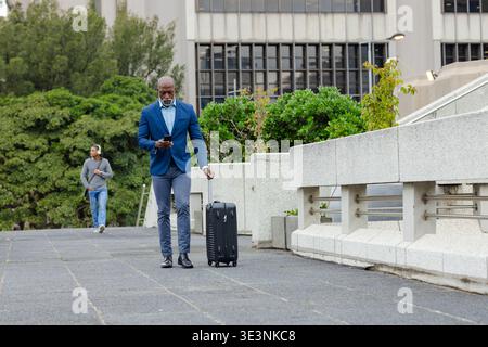 Homme afro-américain en tenue d'affaires Walking plaza tirant valise vérifiant smartphone Banque D'Images