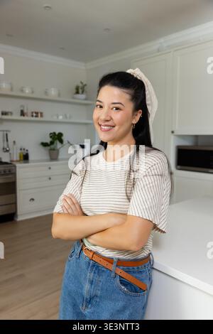 Femme appuyée contre l'îlot de comptoir blanc, pliant les bras dans la cuisine moderne près du micro-ondes Banque D'Images