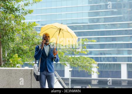 Homme afro-américain mature en costume tenant un parapluie jaune et parlant sur smartphone Banque D'Images