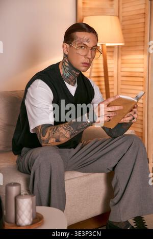 Un jeune homme avec des tatouages et des lunettes aime lire un livre assis dans un cadre élégant et chaleureux. Banque D'Images