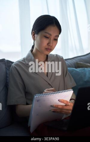Jeune femme adulte asiatique assise sur le canapé écrivant dans un ordinateur portable tout en regardant un ordinateur portable, une expression focalisée, de longs cheveux foncés, participant à l'apprentissage à distance ou travaillant à la maison Banque D'Images