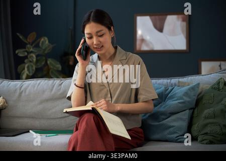 Jeune femme asiatique assise sur un canapé parlant sur un smartphone tout en écrivant dans un ordinateur portable, souriant et se concentrant sur le multitâche, intérieur de la maison avec des éléments décoratifs en arrière-plan Banque D'Images