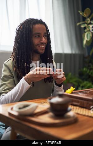 Jeune homme noir adulte assis à une table en bois participant à la cérémonie du thé asiatique traditionnelle tenant une petite tasse de thé souriante s'engageant dans une expérience culturelle à l'intérieur Banque D'Images