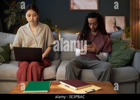 Jeune femme adulte asiatique utilisant un ordinateur portable assis sur un canapé à côté de Black jeune homme adulte naviguant smartphone, les deux axés sur les appareils numériques dans le cadre moderne du salon Banque D'Images