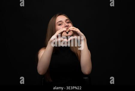 Jeune femme formant une forme de coeur avec les mains, souriant et exprimant l'amour, l'affection et les soins sur fond noir Banque D'Images