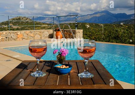 Ambiance relaxante au bord de la piscine dans une villa de vacances en Crète, avec vin rosé, collations et fleurs sur une table en bois donnant sur une piscine privée sereine Banque D'Images