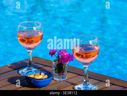 Ambiance relaxante au bord de la piscine dans une villa de vacances en Crète, avec vin rosé, collations et fleurs sur une table en bois donnant sur une piscine privée sereine Banque D'Images