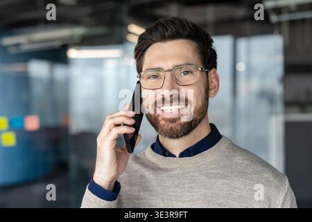 Homme heureux portant des lunettes et un pull ayant un appel téléphonique, souriant tout en communiquant dans un bureau moderne, représentant les affaires, la connexion et le succès Banque D'Images