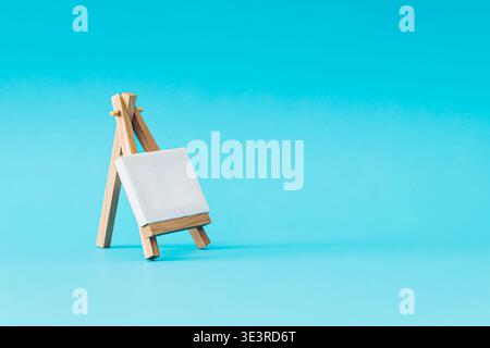 Toile vierge sur chevalet en bois sur fond bleu clair. Maquette artistique minimale. Idée de présentation de marque. Espace de copie créatif. Banque D'Images