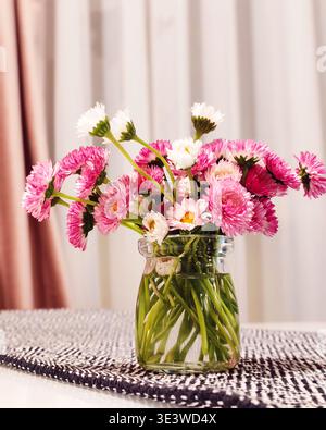 Petit bouquet de fleurs roses et blanches placé dans un bocal en verre transparent sur une table avec un chemin à motifs. Le moment intérieur calme met en valeur la maison Banque D'Images