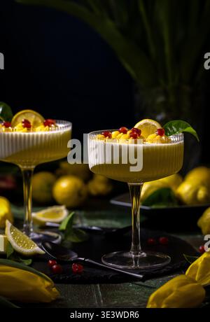 Deux desserts à la panna cotta au citron dans des verres élégants, garnis de graines de grenade, de fruits coupés en dés et de tranches de citron, présentés sur une table sombre Banque D'Images