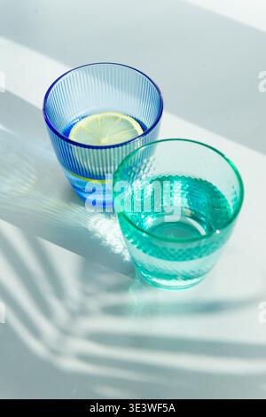 Deux verres texturés, l'un bleu avec une tranche de citron flottant dans l'eau, et l'autre vert, reposent élégamment sur une surface ensoleillée, projetant des ombres complexes Banque D'Images