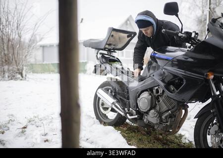 Personne inspectant une moto dans une cour enneigée près d'une maison Banque D'Images