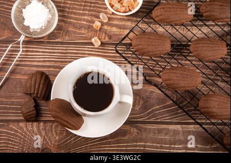 Des madeleines en chocolat en forme de coquille sont exposées avec une tasse de café noir sur une table en bois rustique. Un tamis avec du sucre est à proximité, ajoutant charme t Banque D'Images