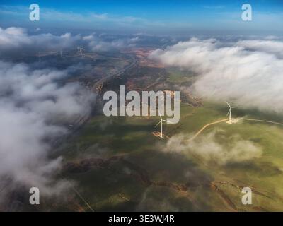 Vue aérienne capturant des éoliennes sur un paysage luxuriant, partiellement entouré de nuages moelleux. La scène illustre l'harmonie de l'énergie renouvelable an Banque D'Images
