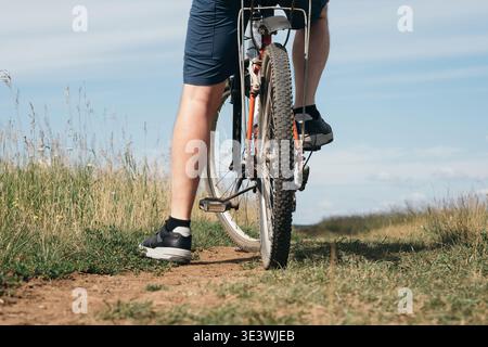 Un cycliste portant un short roule sur un VTT robuste le long d'un chemin de terre. Le chemin est bordé de hautes herbes sous un ciel bleu clair, capturant un moment Banque D'Images