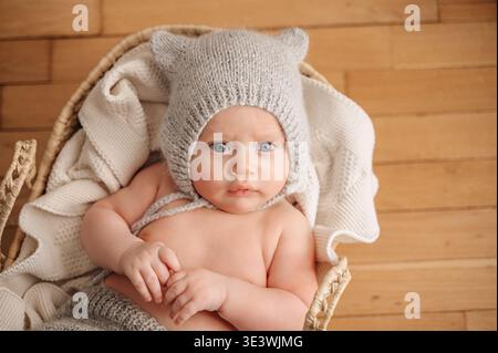Un bébé de 3 mois portant un joli chapeau tricoté avec de petites oreilles se trouve paisiblement dans un panier tissé confortable sur un plancher en bois, entouré de blanke crème douce Banque D'Images