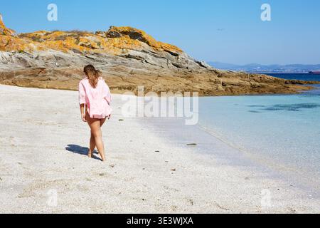 Une femme en chemise rose marche le long de la plage de sable immaculée des îles Cies en Galice, Espagne, avec une eau bleu clair, des affleurements rocheux et lointains Banque D'Images