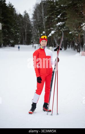 Un skieur vêtu d'une tenue d'hiver rouge vif se tient confiant sur un sentier enneigé en forêt. Tenant des bâtons de ski à la main, ils sont entourés par de grands evergr Banque D'Images