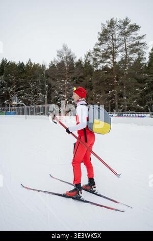 Skieur de fond avec tenue rouge et sac à dos glissant à travers un sentier enneigé pittoresque entouré d'arbres d'hiver, mettant en valeur le plaisir et Banque D'Images
