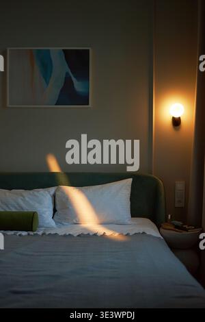 Vertical de chambre moderne vide montrant un lit soigneusement fait avec des oreillers, peinture abstraite sur le mur, applique murale illuminée jetant une lumière chaude, aucune personne visible, scène intérieure tranquille Banque D'Images