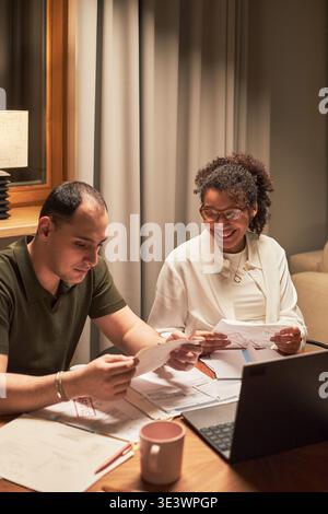 Vertical de l'homme caucasien d'âge moyen examinant des documents avec une femme noire d'âge moyen souriant à côté de lui, tous deux assis à table avec un ordinateur portable et des documents, collaborant sur le projet à l'intérieur Banque D'Images