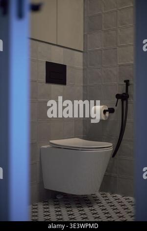Vertical de l'intérieur moderne de salle de bains avec toilettes fixées au mur, porte-papier toilette, plaque de chasse noire et pulvérisateur de bidet à main contre le mur carrelé, sol à motifs géométriques visible Banque D'Images