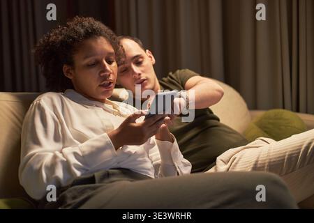 Jeune femme noire et jeune homme caucasien assis sur un canapé à l'aide d'un smartphone ensemble, femme tenant le dispositif tandis que l'homme pointant vers l'écran, tous deux axés sur l'interaction de la technologie mobile Banque D'Images