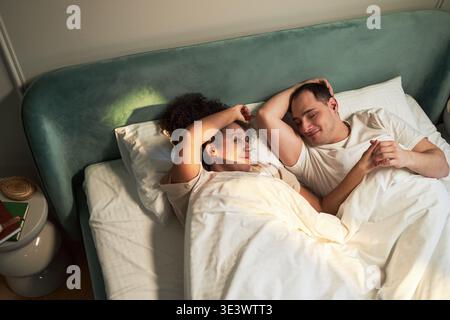 Jeune adulte femme noire et jeune adulte homme caucasien allongé dans le lit ensemble sous une couverture, face à face et souriant, moment du matin détendu dans le cadre de la chambre Banque D'Images