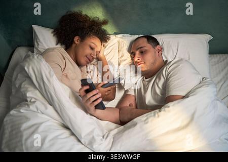 Jeune adulte femme noire et jeune adulte homme caucasien allongé dans le lit à l'aide de smartphones, les deux regardant des écrans tout en se relaxant ensemble sous une couverture, les têtes reposant sur des oreillers Banque D'Images