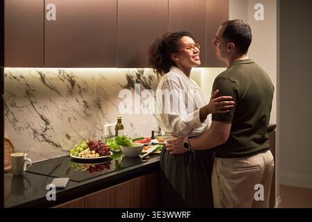 Jeune femme noire adulte et jeune homme caucasien adulte embrassant dans la cuisine moderne, souriant et faisant un contact visuel tout en préparant des fruits et légumes frais sur le comptoir Banque D'Images