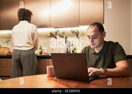 Jeune homme caucasien adulte travaillant sur un ordinateur portable à la table de la cuisine tandis qu'une femme noire d'âge moyen debout au comptoir préparant la nourriture dans l'intérieur de la cuisine moderne Banque D'Images